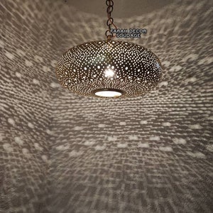 Może przedstawiać: Złota metalowa lampa wisząca z okrągłym, perforowanym kloszem. Lampa jest zawieszona na łańcuchu i rzuca wzorzysty cień na sufit.