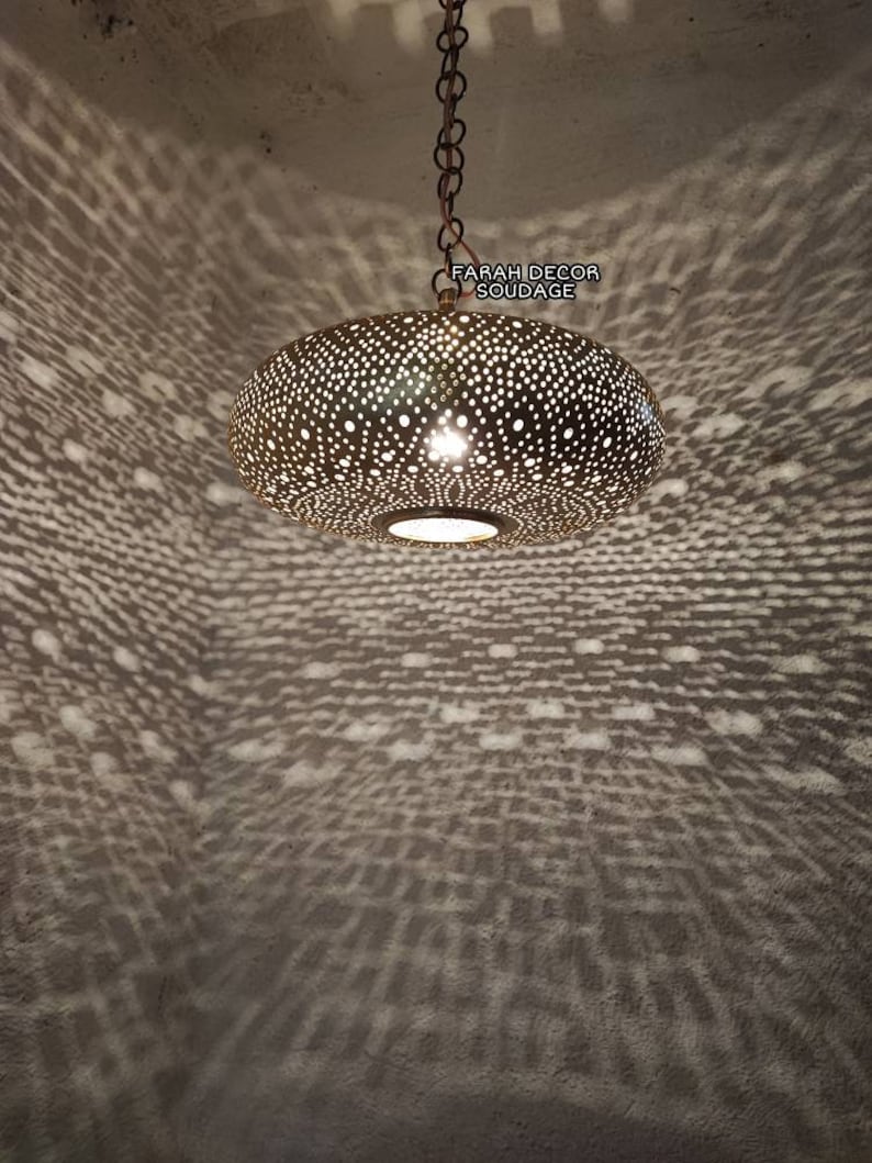 Może przedstawiać: Złota metalowa lampa wisząca z okrągłym, perforowanym kloszem. Lampa jest zawieszona na łańcuchu i rzuca wzorzysty cień na sufit.