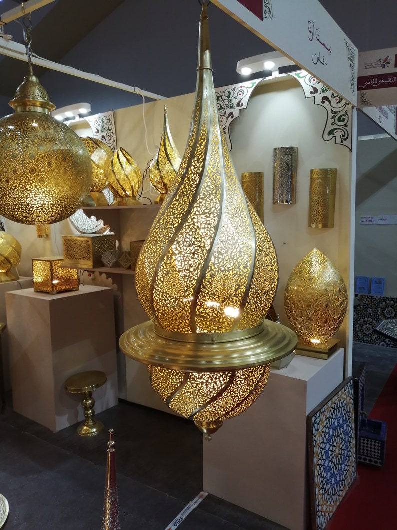 May include: A gold metal hanging lamp with intricate cut-out designs. The lamp has a round top and a teardrop-shaped bottom. The lamp is lit and casting a warm glow.Moroccan Brass Pendant Light - Handmade Chandelier Lighting - Home Ceiling Decor .