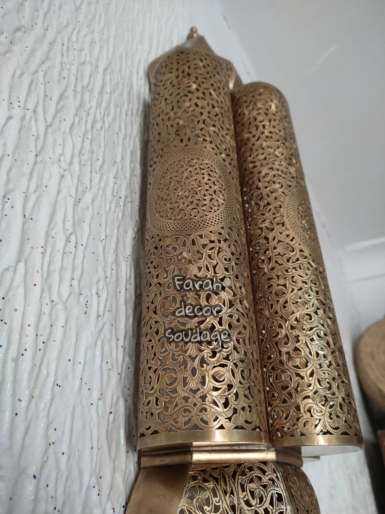 May include: Two gold metal lanterns with intricate cut-out designs. The lanterns are attached to a wall and have a decorative base.