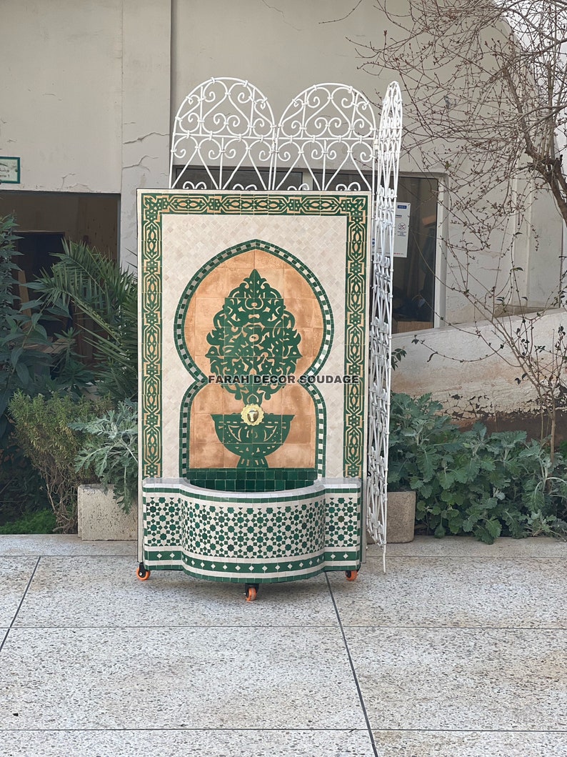 Puede incluir: Una fuente marroqu&iacute; decorativa con azulejos verdes y blancos. La fuente presenta un dise&ntilde;o de arco tradicional y un motivo floral central. La fuente est&aacute; sobre ruedas y tiene el texto "FARAH DECOR SOUADIA" en la parte inferior.