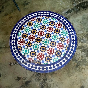Può includere: Un tavolo rotondo con un piano a mosaico caratterizzato da un motivo geometrico di piastrelle blu, gialle, rosse, verdi e bianche. Il bordo presenta un disegno a rombi blu e bianchi. Il testo Farah decor Soudage è visibile sulla superficie.