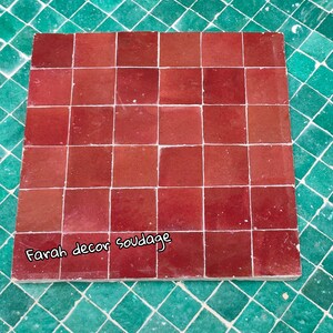 May include: A close-up of a red tile sample with the text "Farah decor soudagge" written on the bottom. The tile sample is made up of 25 individual tiles.