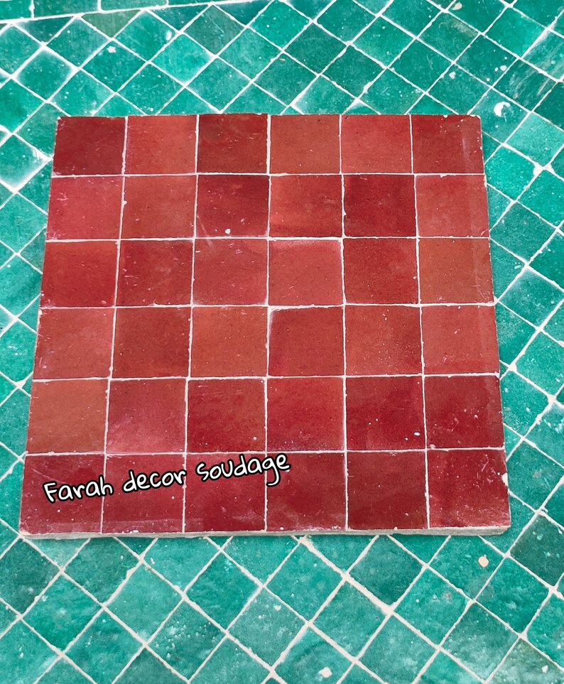 May include: A close-up of a red tile sample with the text "Farah decor soudagge" written on the bottom. The tile sample is made up of 25 individual tiles.