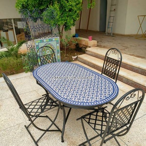 May include: A white and blue mosaic oval table with a black metal frame and four black metal chairs with a criss-cross pattern on the seat. The table and chairs are on a grey tiled patio. The text "FARAH DECOR SOUDAGE" is visible on the table.