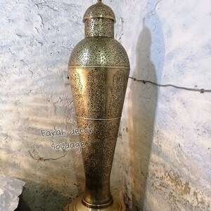 K&ouml;nnte beinhalten: Eine hohe, goldfarbene Metalllampe mit filigranen Ausschnitten. Die Lampe hat einen runden Sockel und einen gew&ouml;lbten oberen Teil. Die Lampe steht auf einem kleinen, quadratischen Sockel.