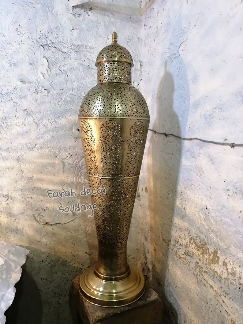 K&ouml;nnte beinhalten: Eine hohe, goldfarbene Metalllampe mit filigranen Ausschnitten. Die Lampe hat einen runden Sockel und einen gew&ouml;lbten oberen Teil. Die Lampe steht auf einem kleinen, quadratischen Sockel.
