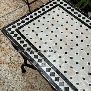 May include: A black and white Moroccan tile table with a geometric pattern. The table has a black metal frame and is sitting on a concrete surface.