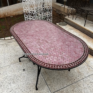May include: A pink and red mosaic tile oval table with a black metal frame. The table is sitting on a gray concrete patio. The text "FARAH DECOR SOUDAGE" is visible on the table.