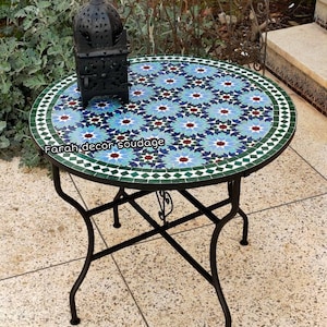 Könnte beinhalten: Ein runder Mosaiktisch mit einem geometrischen Muster in Blau, Grün und Weiß. Der Tisch hat ein schwarzes Metallgestell mit einem dekorativen Rankendesign. Der Tisch steht auf einem hellbraunen Fliesenboden.