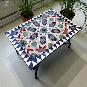 May include: Rectangular coffee table with a mosaic tile top featuring geometric patterns in red, blue, and green. The table has a black metal frame and legs. The border of the table is decorated with blue and white triangles. The text "FARAH DECOR EQUDAE" is visible.