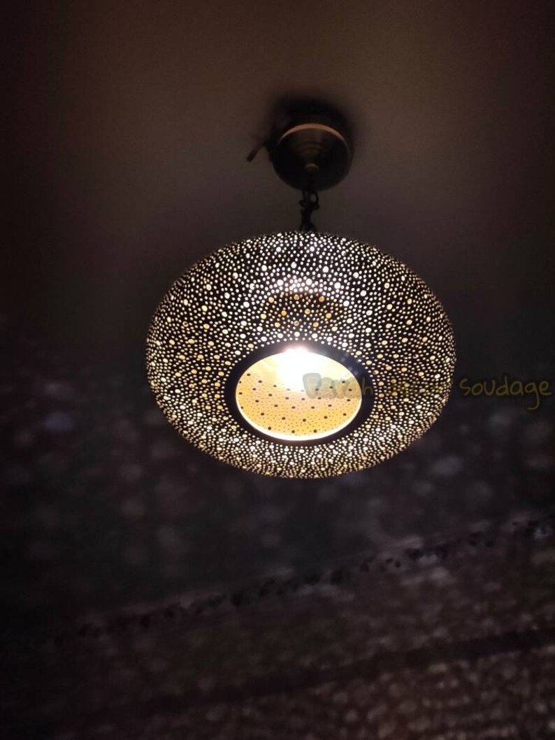May include: A decorative pendant light fixture with a perforated, spherical shade. The shade is dark with numerous small holes, allowing light to filter through. The light source is visible at the bottom, creating a warm glow. The fixture hangs from the ceiling.
