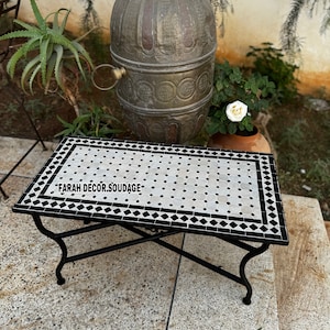 May include: A black metal table with a white and black mosaic tile top. The table has a simple design with a rectangular top and four legs. The mosaic tiles are arranged in a diamond pattern. The table is sitting on a concrete patio.