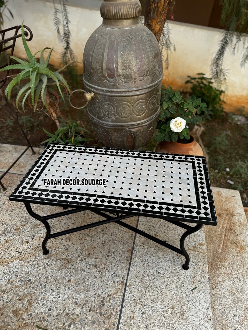 May include: A black metal table with a white and black mosaic tile top. The table has a simple design with a rectangular top and four legs. The mosaic tiles are arranged in a diamond pattern. The table is sitting on a concrete patio.