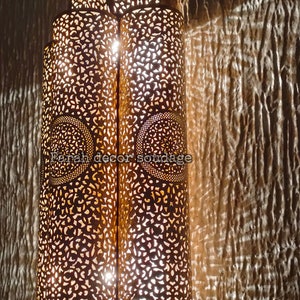 May include: A decorative metal lantern with intricate cut-out designs. The lantern is illuminated from within, casting a warm glow. The text "Farah decor soudaqe" is visible on the lantern.