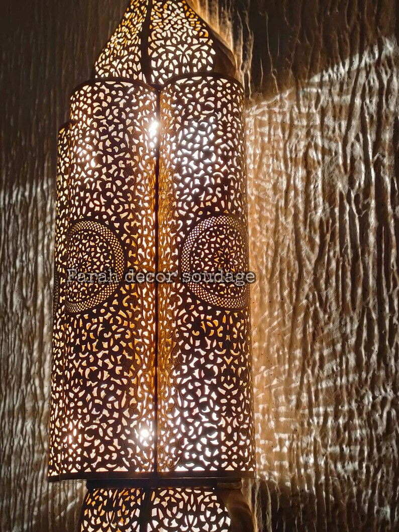 May include: A decorative metal lantern with intricate cut-out designs. The lantern is illuminated from within, casting a warm glow. The text "Farah decor soudaqe" is visible on the lantern.