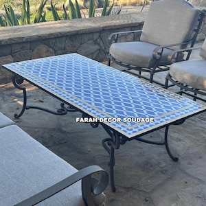 May include: A blue and white tiled coffee table with a black wrought iron frame. The table is sitting on a stone patio with a view of a mountain range in the background. The text "FARAH DECOR SOUDAGE" is visible on the table.