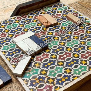 May include: A close-up of a colorful mosaic tile floor with a geometric pattern. The tiles are arranged in a repeating pattern of red, green, blue, yellow, and black. There are also some broken tiles on the floor, and a few pieces of tile are lying on top of the floor. The text "Farah decor soudaqe" is visible on the image.