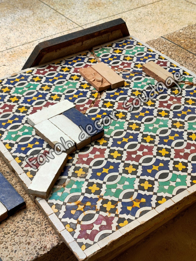 May include: A close-up of a colorful mosaic tile floor with a geometric pattern. The tiles are arranged in a repeating pattern of red, green, blue, yellow, and black. There are also some broken tiles on the floor, and a few pieces of tile are lying on top of the floor. The text "Farah decor soudaqe" is visible on the image.
