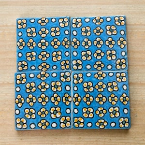 May include: Four square ceramic tiles with a blue background and a repeating floral pattern in yellow, white, and green. The tiles are arranged together on a light brown wooden surface.