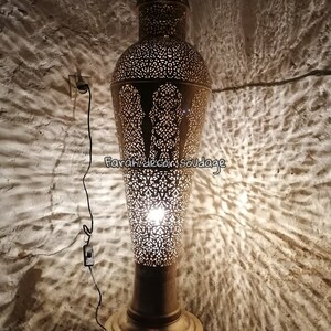 K&ouml;nnte beinhalten: Eine goldene Stehlampe aus Metall mit filigranen Ausschnitten und einer Gl&uuml;hbirne, die das Design beleuchtet. Die Lampe steht auf einer Holzoberfl&auml;che und wirft Schatten an die Wand.