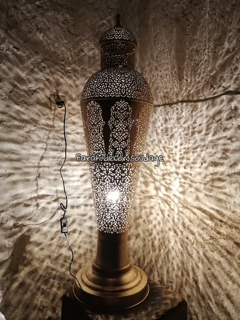 K&ouml;nnte beinhalten: Eine goldene Stehlampe aus Metall mit filigranen Ausschnitten und einer Gl&uuml;hbirne, die das Design beleuchtet. Die Lampe steht auf einer Holzoberfl&auml;che und wirft Schatten an die Wand.