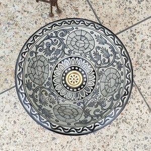 May include: A white ceramic sink with a black and grey floral design. The sink has a gold drain cover and a black rim.