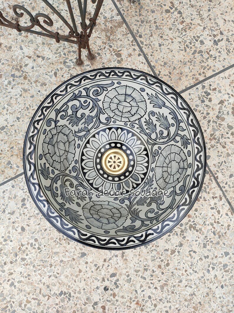 May include: A white ceramic sink with a black and grey floral design. The sink has a gold drain cover and a black rim.
