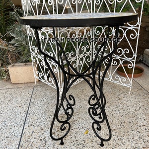 Puede incluir: Una mesa de metal negro con una tapa redonda de mosaico de azulejos blancos y azules. La mesa tiene una base decorativa con volutas y se encuentra sobre una superficie de hormig&oacute;n gris. El texto "FARAH DECOR SOUDAGE" es visible en la mesa.