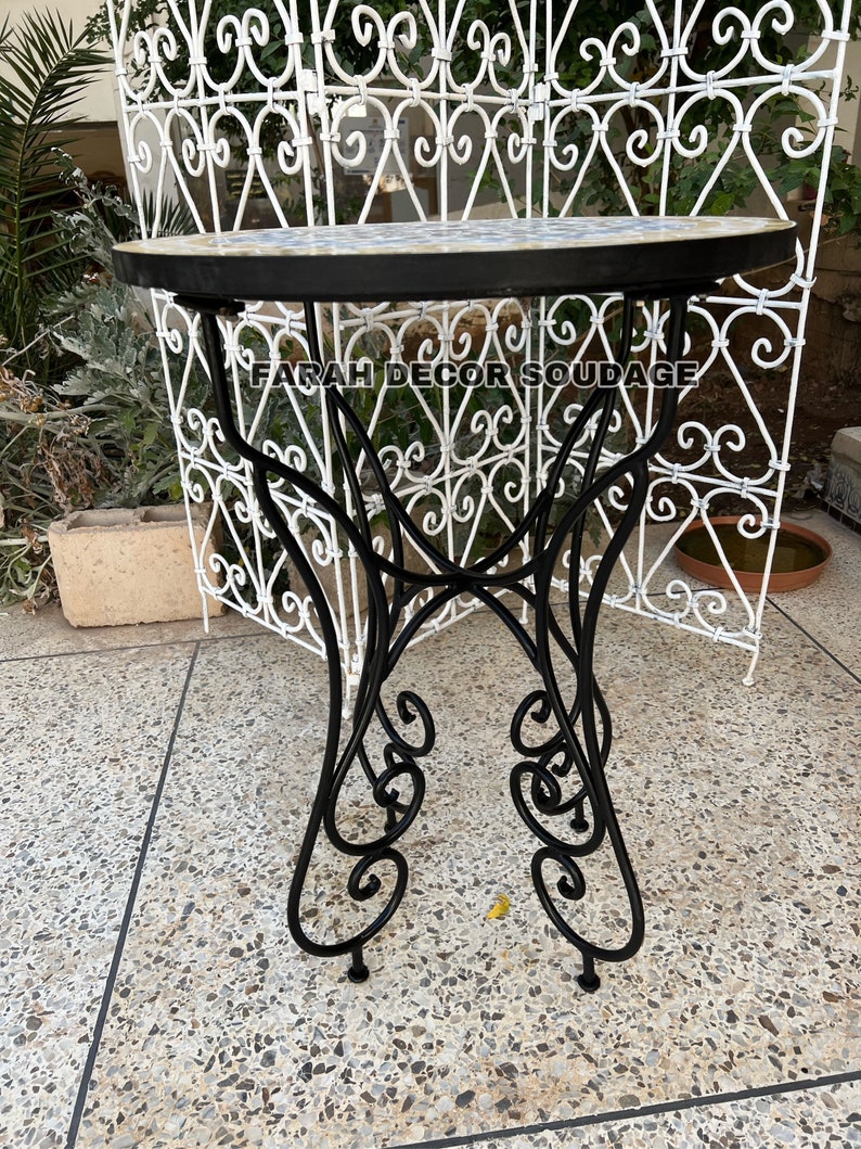 Puede incluir: Una mesa de metal negro con una tapa redonda de mosaico de azulejos blancos y azules. La mesa tiene una base decorativa con volutas y se encuentra sobre una superficie de hormig&oacute;n gris. El texto "FARAH DECOR SOUDAGE" es visible en la mesa.
