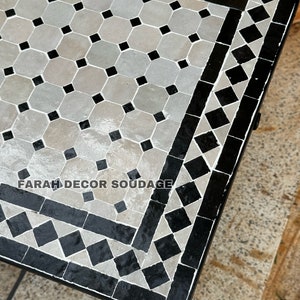 May include: A black and white Moroccan tile table top with a geometric pattern. The table top is made of small, square tiles arranged in a repeating pattern. The table top is surrounded by a black border.