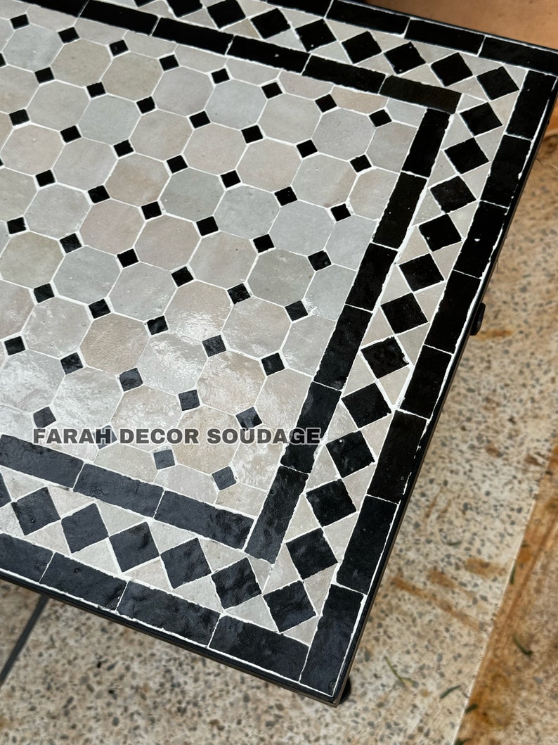 May include: A black and white Moroccan tile table top with a geometric pattern. The table top is made of small, square tiles arranged in a repeating pattern. The table top is surrounded by a black border.