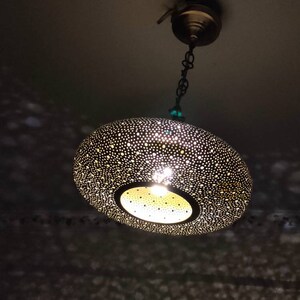 May include: A decorative, hanging pendant light with a perforated, bronze-colored shade. The light fixture is suspended from a chain and ceiling mount. The shade's design casts a pattern of light and shadow.