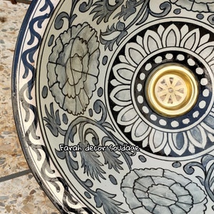 May include: A white ceramic bowl with a black and grey floral design. The bowl has a gold-coloured metal insert in the centre. The text "Farah decor Soudage" is written on the bowl.