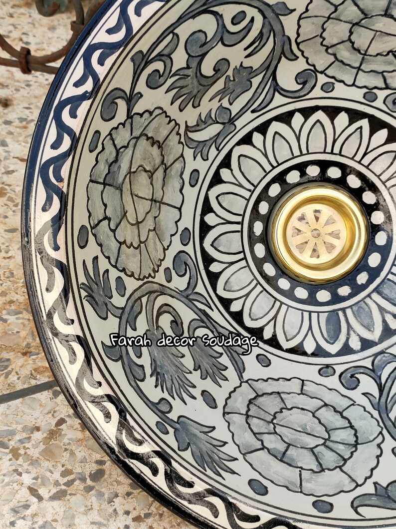 May include: A white ceramic bowl with a black and grey floral design. The bowl has a gold-coloured metal insert in the centre. The text "Farah decor Soudage" is written on the bowl.