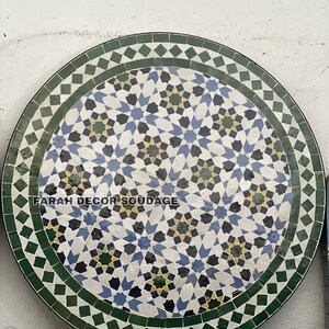 May include: A round, green, white, blue, and yellow mosaic tile table top with a geometric pattern. The table top has a black border and the text "FARAH DECOR SOUDAGE" in the center.