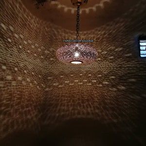 Może przedstawiać: Okrągła, miedziana lampa wisząca w stylu marokańskim z ozdobnym, skomplikowanym wzorem małych otwor&oacute;w. Lampa jest zawieszona na łańcuchu i rzuca geometryczny wz&oacute;r światła na sufit.