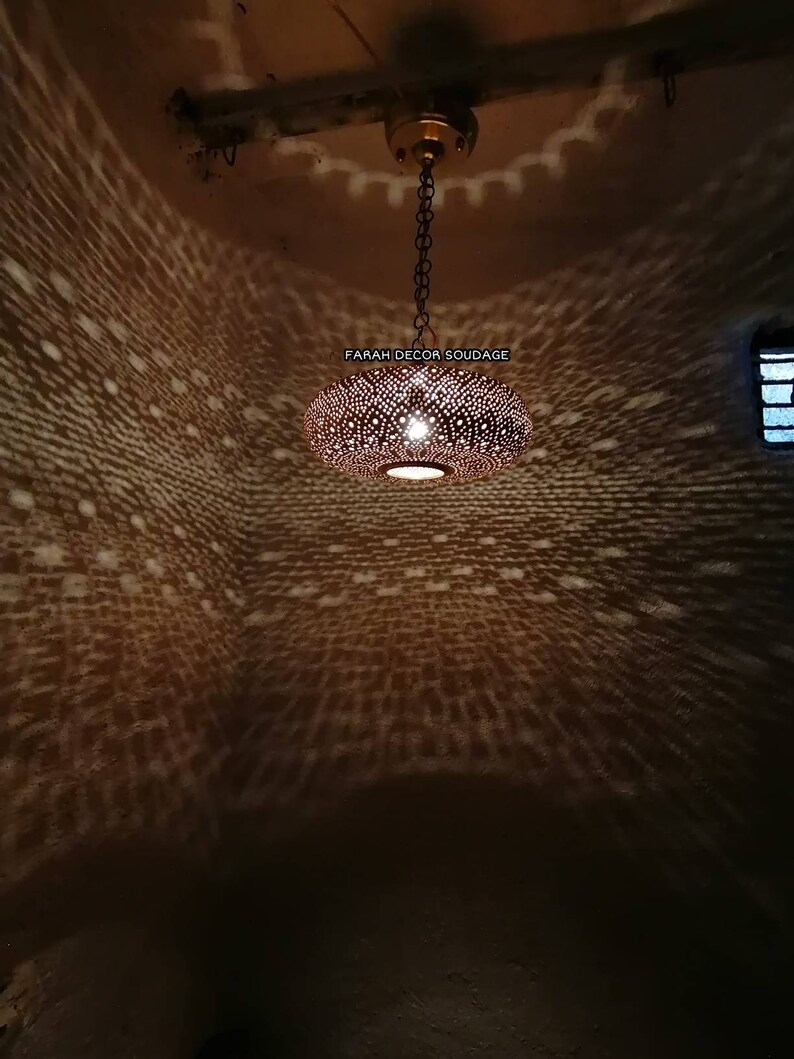 Może przedstawiać: Okrągła, miedziana lampa wisząca w stylu marokańskim z ozdobnym, skomplikowanym wzorem małych otwor&oacute;w. Lampa jest zawieszona na łańcuchu i rzuca geometryczny wz&oacute;r światła na sufit.