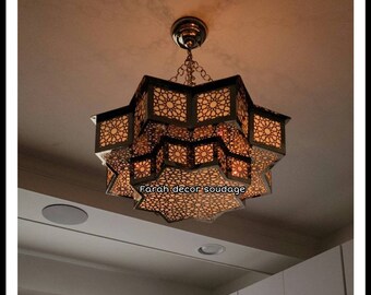 May include: A multi-tiered, star-shaped, metal chandelier with intricate cut-out designs. The chandelier is lit and casts a warm glow. The text "Farah decor soudagé" is visible in the image.