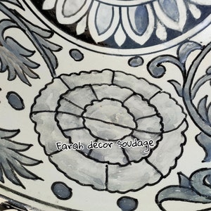 May include: A white ceramic sink with a black and white floral design. The sink is round and has a drain hole in the centre. The design features a large flower in the centre with smaller flowers and leaves surrounding it. The text "Farah decor soudaage" is written in black on the sink.
