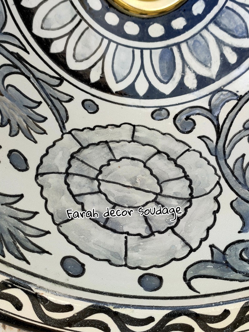 May include: A white ceramic sink with a black and white floral design. The sink is round and has a drain hole in the centre. The design features a large flower in the centre with smaller flowers and leaves surrounding it. The text "Farah decor soudaage" is written in black on the sink.