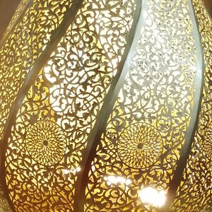 May include: A gold metal Moroccan lantern with intricate cut-out designs. The lantern is illuminated from within, casting a warm glow through the delicate patterns.