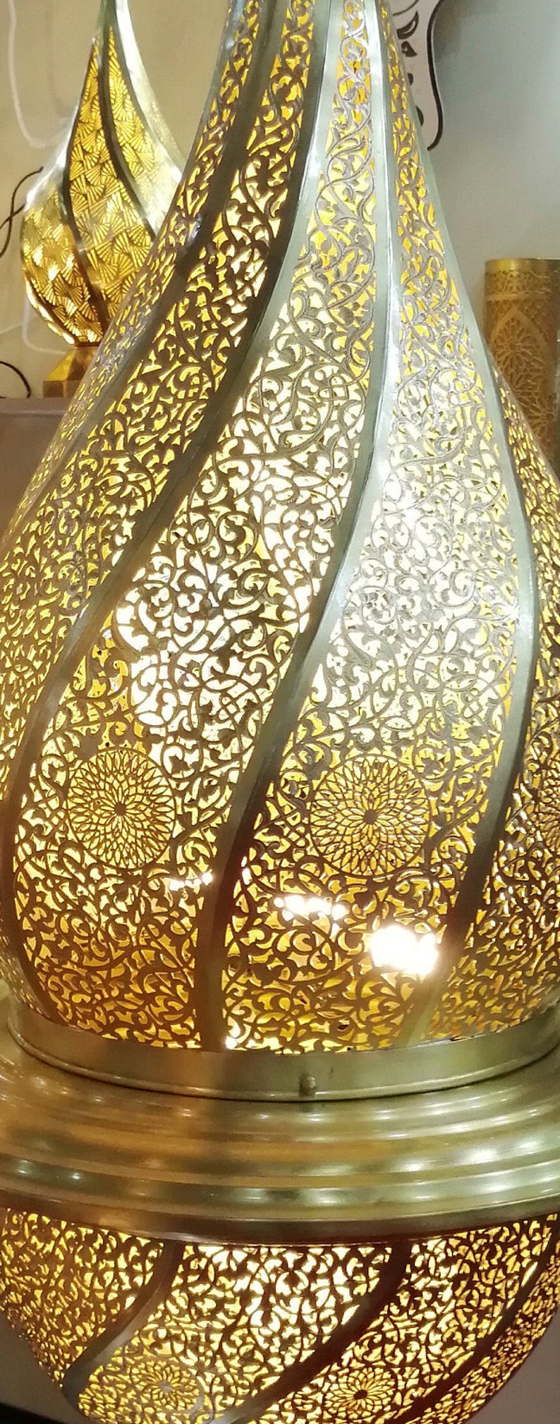 May include: A gold metal Moroccan lantern with intricate cut-out designs. The lantern is illuminated from within, casting a warm glow through the delicate patterns.