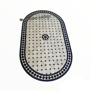 May include: Oval-shaped white and blue mosaic tile table top with a black border. The tile pattern features a repeating diamond design with a small blue and white flower in the centre.