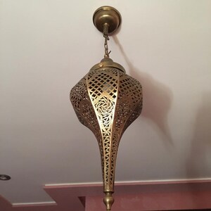 May include: A gold metal pendant light fixture with intricate cut-out designs. The light fixture is hanging from a chain and has a pointed bottom.