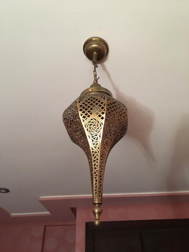 May include: A gold metal pendant light fixture with intricate cut-out designs. The light fixture is hanging from a chain and has a pointed bottom.