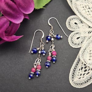 May include: A pair of dangle earrings featuring silver-toned wire hooks and multiple strands of faceted beads. The beads are a mix of deep blue and magenta pink, accented with small silver-toned metal spacers, creating a layered, cascading design.
