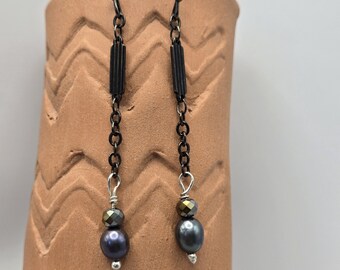 May include: A pair of dangle earrings with a dark gunmetal finish. Each earring features a chain, a rectangular bar, a faceted bead, and a dark gray pearl-shaped bead. The earrings are displayed against a textured, brown background.
