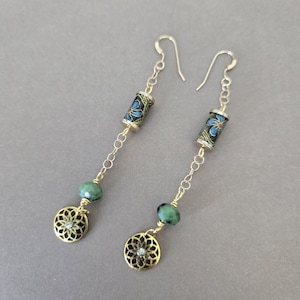 May include: A pair of dangle earrings with gold-colored chains and accents. Each earring features a cylindrical bead with a blue floral design, a green bead, and a gold-colored filigree charm. The earrings hang from silver-colored ear wires.