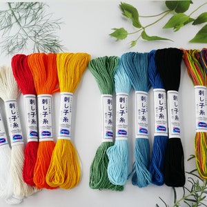 May include: A selection of embroidery thread skeins in a range of colours: white, red, orange, yellow, green, blue, and black. Each skein is labelled with Japanese text. Needles and foliage are also visible.